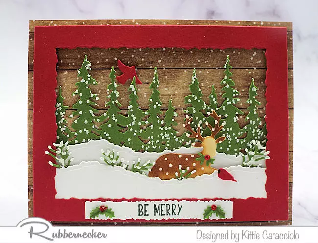 A beautiful handmade winter scene Christmas card set in a red frame with a woodgrain background showing snowy evergreens and a docile deer.