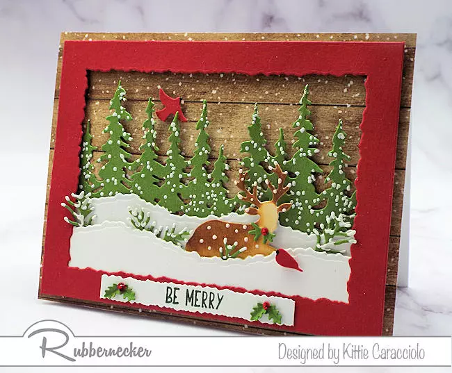A handmade winter scene Christmas card with a deer among snowbanks set against a row of evergreens.