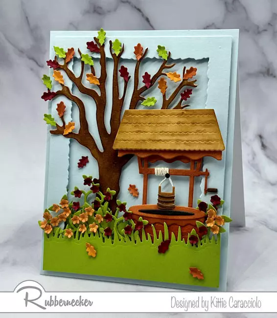 A card with a die cut wishing well set in an Fall scene that is so realistic thanks to detailed dies from Rubbernecker used to make it.