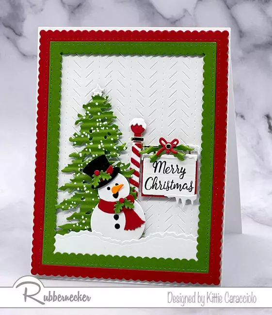 An adorable handmade die cut snowman card with details like a snowy tree, a striped sign pole and a textured background.