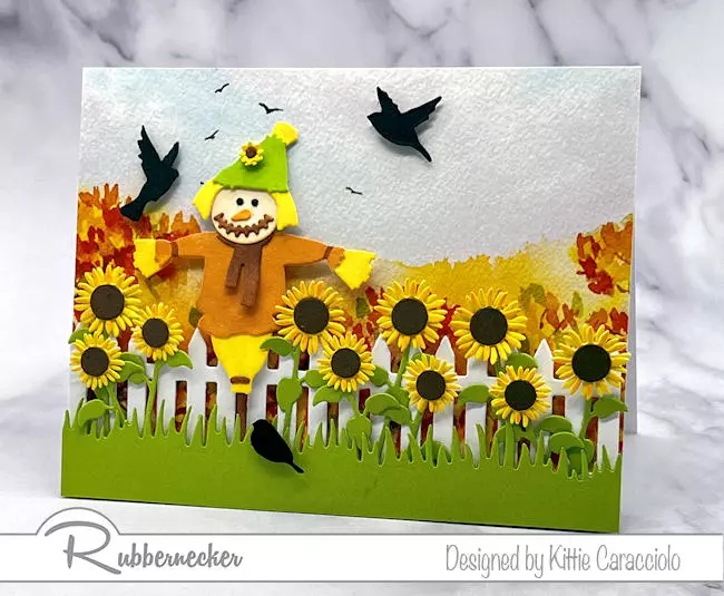 A cute handmade scarecrow card made using a preprinted watercolor look background paper and decorative die cuts, all from Rubbernecker.