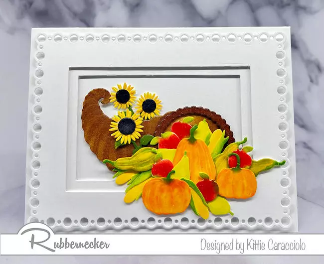 Die cut and hand colored fruits and veggies spilling from a cornucopia are set in a beautiful white on white layered rectangle die cut frame on today